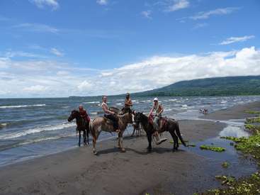 The image depicts four individuals riding horses on a beach, with a serene ocean and mountainous backdrop under a blue sky with white clouds.