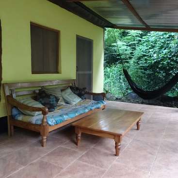 The image depicts a serene outdoor patio with a wooden bench, coffee table, and hammock, surrounded by lush greenery and a vibrant yellow wall.