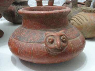 The image features an ancient clay pot with a wide mouth and narrow neck, adorned with a face on the side, surrounded by other similar artifacts on a white surface.
