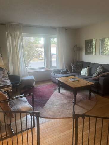 Un salon chaleureux avec de grandes fenêtres, des rideaux blancs, un canapé d'angle gris, un tapis rond, une table basse en bois, un fauteuil, des décorations murales et une barrière de sécurité pour bébé.