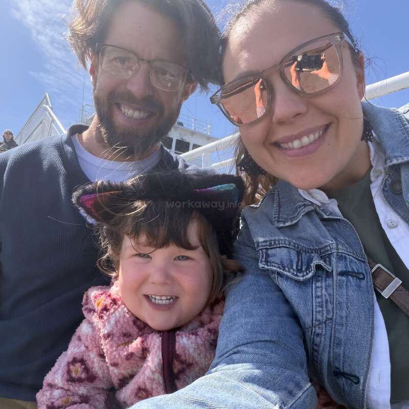 Une famille souriante profite d'une journée ensoleillée à l'extérieur. Le père porte des lunettes, la mère des lunettes de soleil, et leur fille un manteau rose et un bandeau à oreilles de chat.