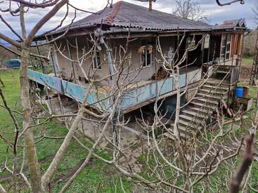 An old, weathered house stands elevated on stilts, surrounded by leafless branches and green grass. It has a balcony, stairs, and a rustic rural atmosphere.