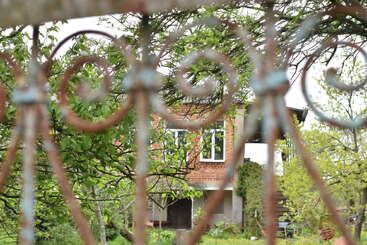 A rustic house with red bricks and white windows stands behind an ornate, rusted metal fence, surrounded by lush green trees and plants in springtime.