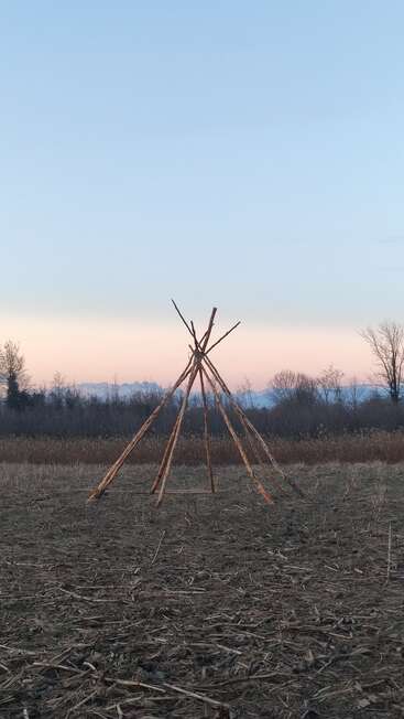 A simple wooden structure made of logs stands in an open field at dusk, with barren ground, distant trees, and a pastel sunset sky behind.