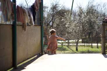A young child in a dress stands on a sunlit porch, reaching outward. Laundry hangs above, and bare trees and greenery fill the bright background.