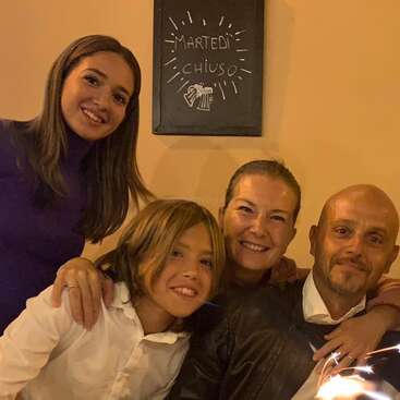 The image shows a family of four posing for a photo, with a sign on the wall behind them that appears to be written in Italian.