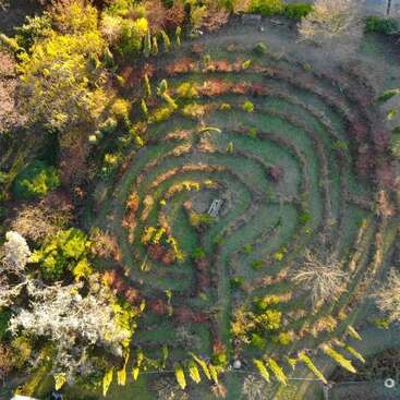 A imagem mostra uma vista aérea de um jardim sereno com um grande e intrincado padrão em espiral de grama e plantas, cercado por árvores e arbustos.
