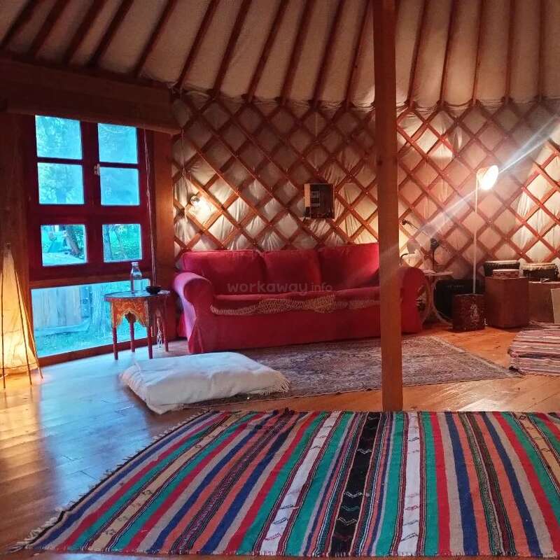A imagem mostra o interior de uma yurt aconchegante com um sofá vermelho, tapete colorido e piso de madeira, com uma grande janela e uma pequena mesa, exalando uma atmosfera calorosa e convidativa.