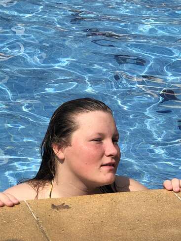 La imagen muestra a una joven con el pelo mojado, de pie en una piscina, con un bañador negro y las manos apoyadas en el borde de la piscina.
