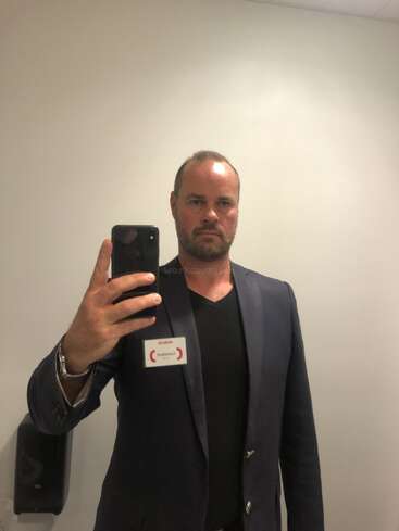 La imagen muestra a un hombre haciéndose un selfie en un espejo, vestido con una chaqueta de traje oscura sobre una camisa negra y sosteniendo un teléfono con una etiqueta con su nombre alrededor del cuello.