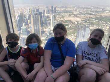 La imagen muestra a cuatro niños sentados frente a una ventana con vistas a la ciudad, todos con máscaras y mirando a la cámara.