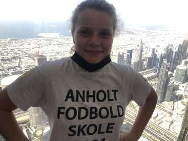 La imagen muestra a una joven delante de un paisaje urbano, con una camiseta blanca y un texto negro que dice "ANHOLT FODBOLD SKOLE".