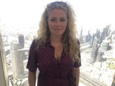 La imagen muestra a una mujer de pie delante de un paisaje urbano, con una marca de agua en la que se lee "workaway.info" sobre el pecho. Lleva el pelo largo y rubio y un vestido morado.