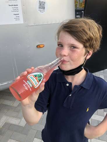 La imagen muestra a un niño con pecas y pelo castaño, vestido con un polo azul marino, bebiendo de una botella de refresco Jarritos Guayaba.