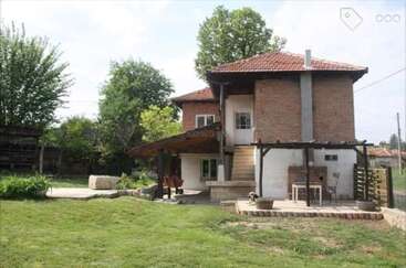 This image shows a rustic two-story brick house with a red-tiled roof, surrounded by greenery, featuring a spacious yard, patio area, and outdoor seating.