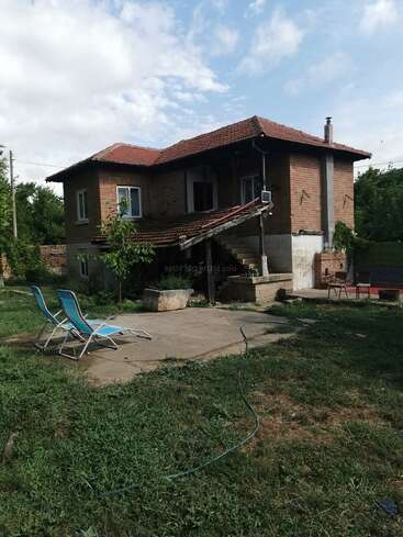 This image shows a rustic two-story brick house with a red tiled roof, outdoor stairs, two blue lounge chairs, green lawn, and partially cloudy sky.