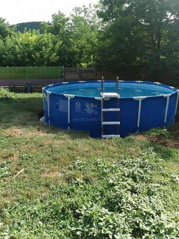 A round blue above-ground pool with a metal ladder sits in a grassy backyard. Trees and a wooden fence surround the area, providing shade and privacy.