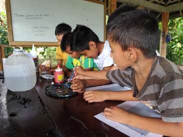 Four young boys sit at a wooden table, concentrating on painting together. Art supplies, water, and paints are on the table. A whiteboard is behind them.