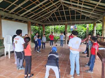 A group of children and a few adults stand in a circle under an open pavilion with a tiled floor, surrounded by lush greenery, engaged in an activity.