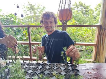 A man smiles while sitting at a table, planting seedlings in soil-filled trays on a porch surrounded by lush greenery, with another person assisting beside him.