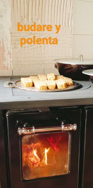 Ein Holzofen mit einem brennenden Feuer, ein Metalltablett mit Polenta-Brotstücken drauf, Töpfe im Hintergrund und der Text „Budare y Polenta”.