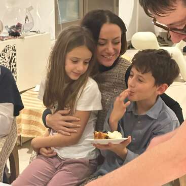 A smiling woman hugs a girl while a boy enjoys dessert, surrounded by friends or family in a cozy, warmly lit indoor setting, creating a joyful moment.