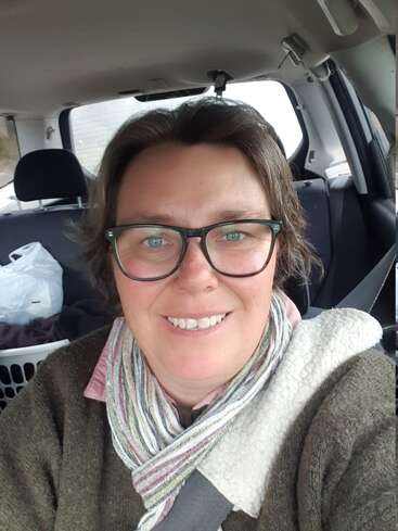Uma pessoa sorridente de óculos está sentada em um carro, usando um cachecol e um suéter marrom. Um cesto de roupa suja e sacolas estão no banco de trás.
