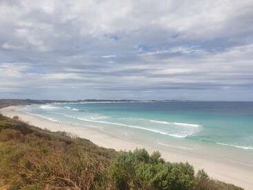 Une superbe scène de plage avec des vagues turquoise s'écrasant doucement sur le sable blanc, encadrées par la végétation côtière. Le ciel est partiellement nuageux, ce qui ajoute à la tranquillité de la scène.