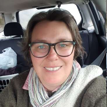 Uma pessoa sorridente de óculos está sentada em um carro, usando um cachecol e um suéter marrom. Um cesto de roupa suja e sacolas estão no banco de trás.