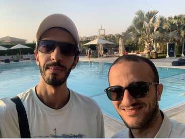 Dois homens de óculos escuros posam para uma foto à beira de uma piscina. Palmeiras, guarda-sóis e um céu azul sugerem um clima de férias relaxante e ensolarado.