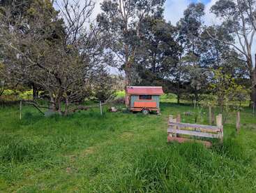 A small mobile shed with a red roof sits on lush green grass, surrounded by trees and a rustic wooden fence, creating a serene rural scene.
