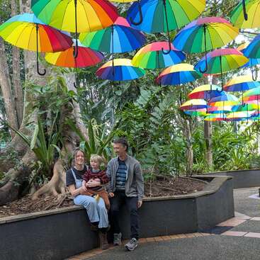 Uma família está sentada em um banco curvo em um jardim exuberante, sorrindo. Guarda-chuvas coloridos estão pendurados no alto, criando uma atmosfera vibrante e alegre em meio à vegetação.