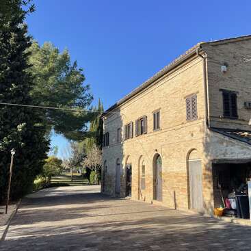 A rustic brick building with arched doorways and shuttered windows sits under a clear blue sky, surrounded by trees and a peaceful stone courtyard.