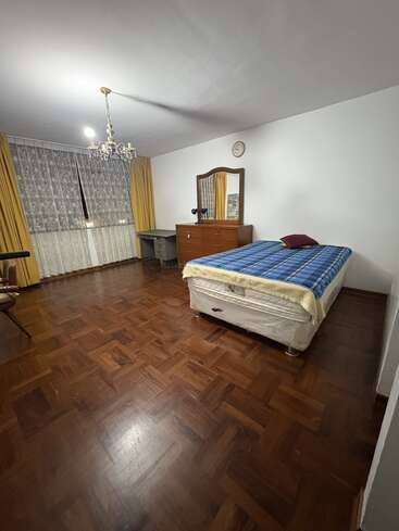 Dies ist ein geräumiges Schlafzimmer mit Holzfußboden, einem Einzelbett, einer Kommode mit Spiegel, einem Schreibtisch, einem Stuhl, gelben Vorhängen, Spitzenvorhängen, einem Kronleuchter und einer Wanduhr.