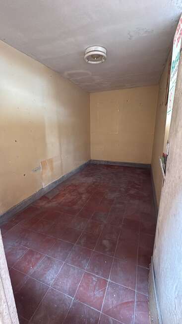 Dies ist ein kleiner, leerer Raum mit gelblichen Wänden, rotem Kachelboden und einer Deckenlampe. Er wirkt abgenutzt, staubig und renovierungsbedürftig.