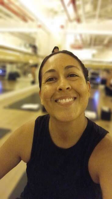 Eine Frau lächelt strahlend und macht ein Selfie in einem Fitnessstudio. Sie trägt ein ärmelloses schwarzes Oberteil. Im Hintergrund sind Trainingsmatten und Spiegel zu sehen.