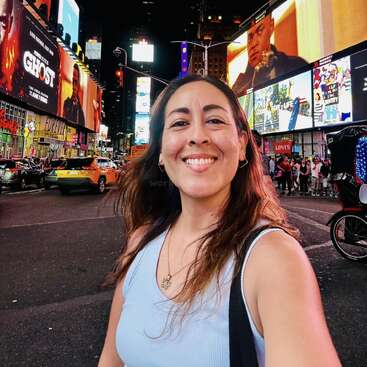 Uma mulher sorridente tira uma selfie à noite na Times Square, em Nova York. Outdoors luminosos, táxis e multidões agitadas compõem o cenário vibrante e colorido.