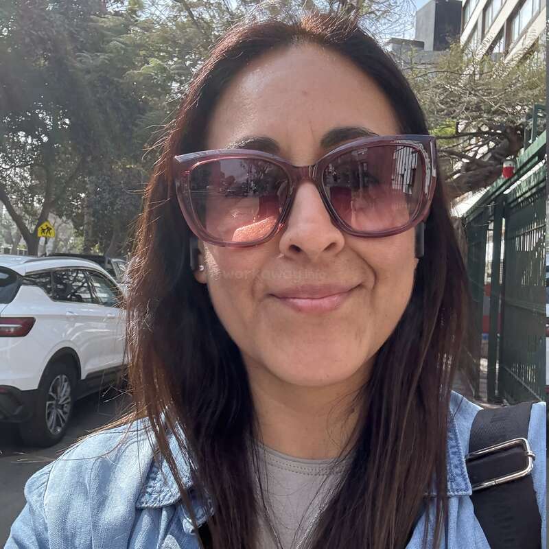 Eine Frau mit langen dunklen Haaren und großer Sonnenbrille lächelt im Freien. Sie trägt eine Jeansjacke, einen Rucksackgurt und kabellose Ohrstöpsel; hinter ihr stehen Bäume und Autos.