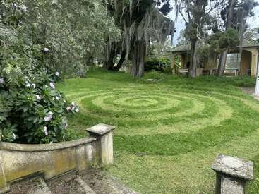 In einem begrünten Garten ist ein spiralförmiges Muster in den Rasen gemäht. Steinstufen und eine Mauer führen zu alten Bäumen, blühenden Sträuchern und einem gelben Gebäude.