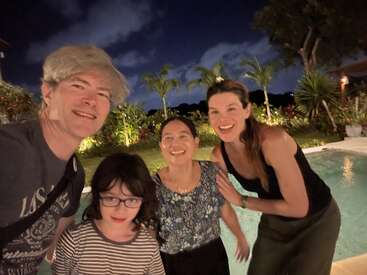 Quatro pessoas, três adultos e uma criança, sorriem felizes à beira de uma piscina à noite. Palmeiras, vegetação exuberante e um céu escuro e nublado criam um cenário tropical.