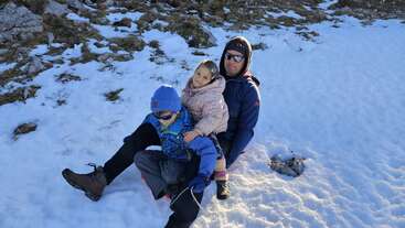 A man and two children, dressed warmly, sit together on snowy ground. They appear to be enjoying a winter outing surrounded by patches of snow and rocky terrain.