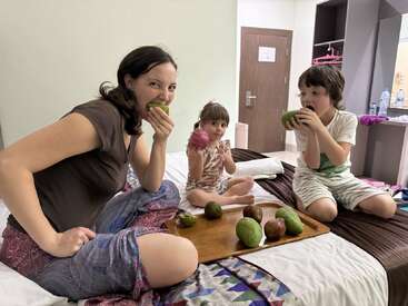 A woman and two children sit on a bed, joyfully eating tropical fruits. A wooden tray with fruits is between them in a cozy, modern room.