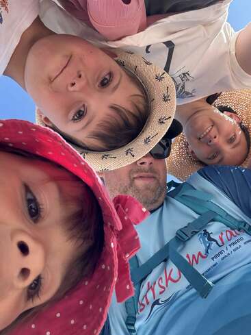 Four people, including two children in hats, lean in close together and smile for a selfie under bright sunlight, creating a playful, joyful family moment.