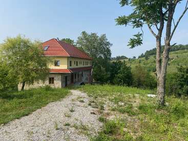 Una encantadora casa con tejado rojo se alza entre exuberante vegetación y colinas onduladas, rodeada de árboles y un camino de grava bajo un cielo azul despejado.
