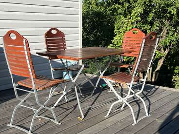 Una mesa de madera para exteriores con cuatro sillas a juego en una terraza junto a una casa, con la luz del sol proyectando sombras y árboles verdes al fondo. Un entorno tranquilo.
