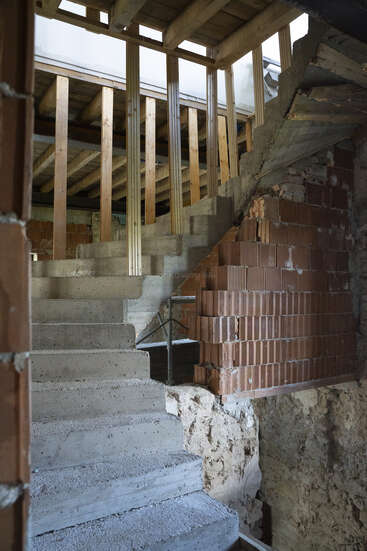 Esta imagen muestra el interior de un edificio inacabado con escaleras de hormigón en bruto, ladrillos rojos a la vista, vigas de madera y estructuras parciales en las paredes, lo que indica una construcción o renovación en curso.