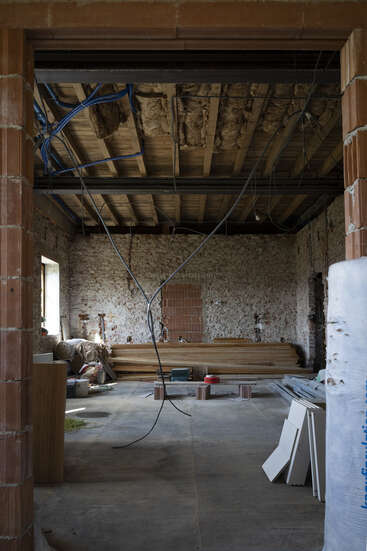La imagen muestra una habitación en construcción, con paredes de ladrillo visto, cables colgando, vigas de madera, materiales de construcción, aislamiento y herramientas esparcidas por el suelo inacabado.