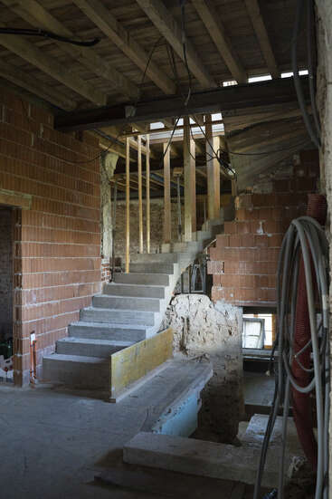Esta imagen muestra un espacio interior inacabado en construcción, con paredes de ladrillo visto, escaleras de hormigón, vigas de madera, cableado visible y diversos materiales de construcción esparcidos.
