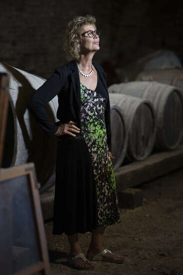 Una mujer con el pelo rizado, gafas y un vestido de flores se alza confiada en una oscura bodega con grandes barriles de madera, iluminada por una suave luz.