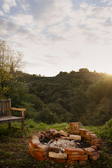 Un banco rústico de madera junto a una hoguera de ladrillo sobre hierba, con vistas a un exuberante valle verde con colinas al atardecer bajo un cielo parcialmente nublado.